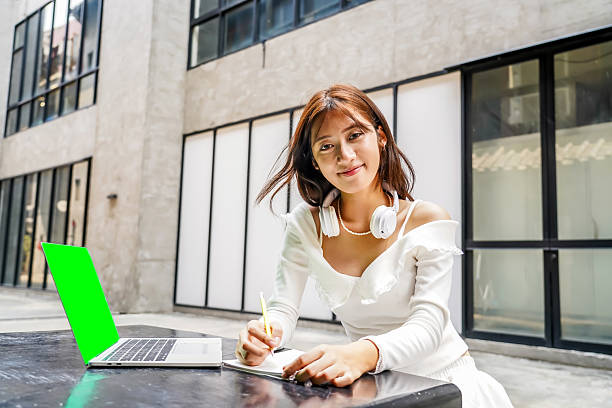 Young asian girl sits outdoors, working on her laptop with a smile. She looks engaged as she takes notes, surrounded by modern architecture and a bright atmosphere, smile friendly