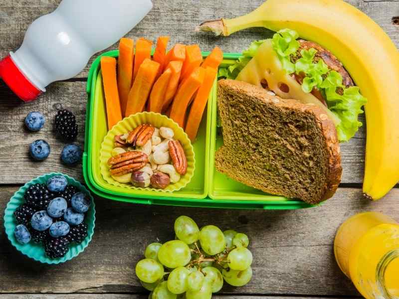 Open lunchbox containing carrots, nuts, sandwich, grapes, and other fresh foods.