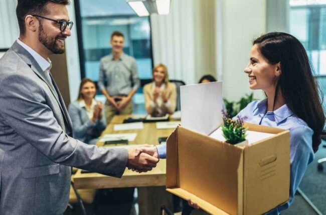 Manager shaking hands with a new employee holding a box on her first day at work, symbolizing onboarding and professional integration.