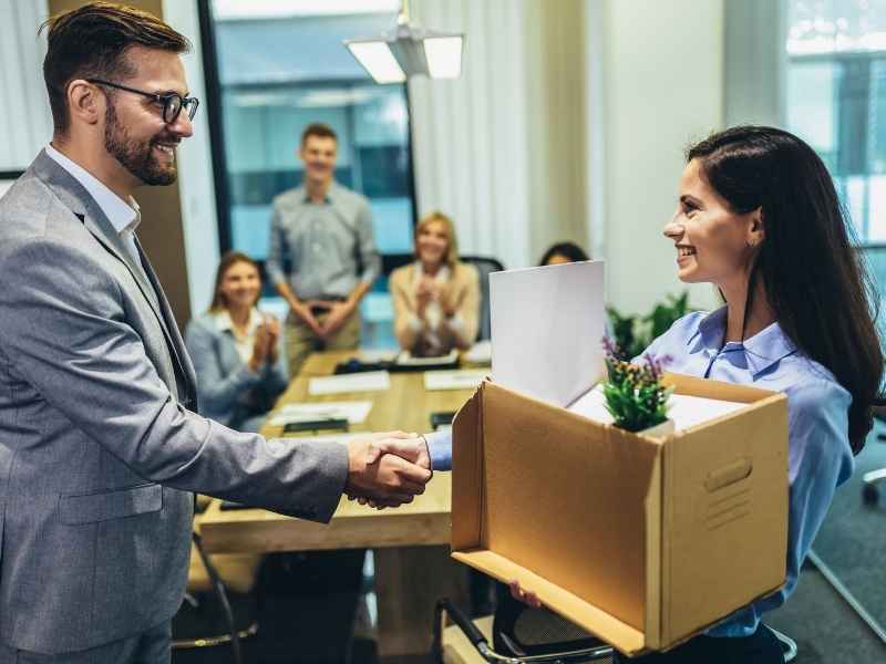 Manager shaking hands with a new employee holding a box on her first day at work, symbolizing onboarding and professional integration.
