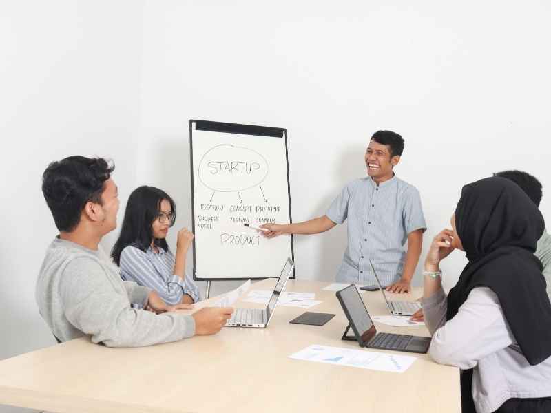 Group of young professionals discussing a startup plan in a meeting room with laptops and a whiteboard, symbolizing teamwork and strategy alignment.