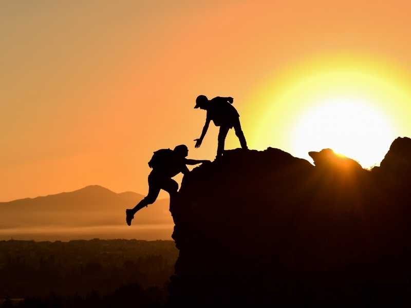 Person helping another climb a rocky peak at sunrise, symbolizing trust and support in business.