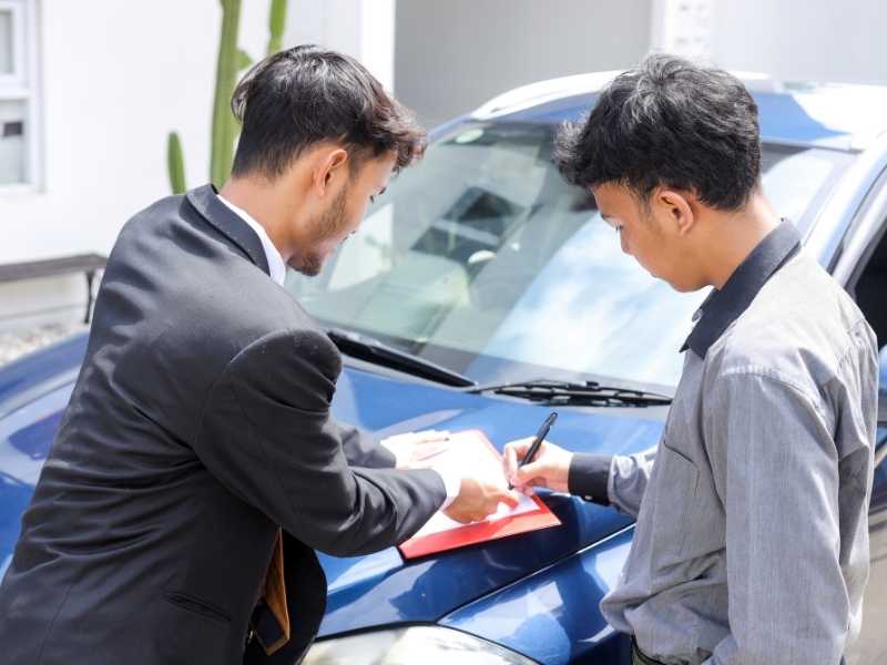 Car owner discussing coverage options with an insurance agent while reviewing documents near a vehicle.