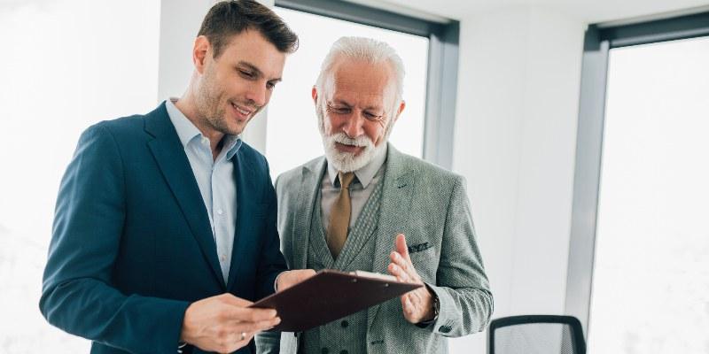 Senior business executive mentoring a younger professional while reviewing documents and discussing leadership responsibilities.